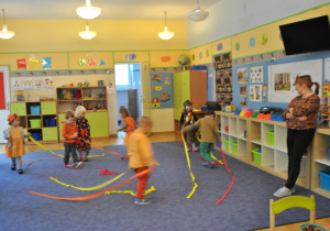 Dzieci tańczą w kole trzymając długie wstążki z kolorowej bibuły. Z prawej strony stoi nauczycielka. Ujęcie 1